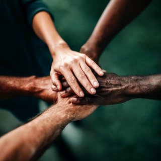 Fünf Hände verschiedener Hauttöne liegen übereinander, was Gemeinschaft und Zusammenarbeit symbolisiert. Die Hände stehen für Einheit und solidarisches Miteinander in einem respektvollen Kontext. Der Hintergrund ist unscharf und dunkel, wodurch der Fokus auf den Händen liegt.
