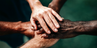 Fünf Hände verschiedener Hauttöne liegen übereinander, was Gemeinschaft und Zusammenarbeit symbolisiert. Die Hände stehen für Einheit und solidarisches Miteinander in einem respektvollen Kontext. Der Hintergrund ist unscharf und dunkel, wodurch der Fokus auf den Händen liegt.