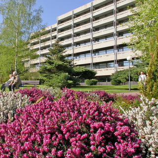 Eine blühende Gartenlandschaft mit rosa und weißen Blumen im Vordergrund. Im Hintergrund ist ein modernes, mehrstöckiges Wohngebäude sichtbar. Einige Personen sitzen und gehen im Garten. Die Szene vermittelt eine ruhige und einladende Atmosphäre.